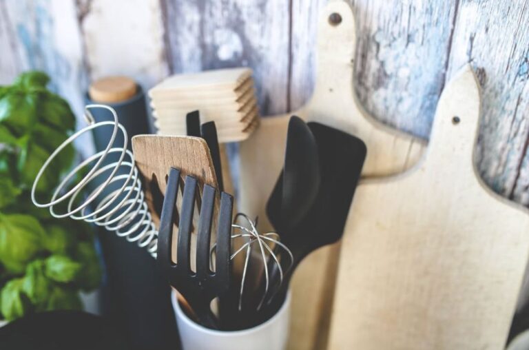 Kitchen utensils in a white holder including whisk, spatula, and fork, with wooden cutting boards and fresh basil in the background.