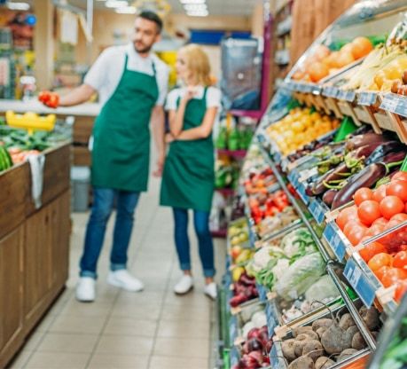Male and female grocery store employees wearing green aprons standing in the fresh produce aisle, surrounded by colorful displays of fruits and vegetables.